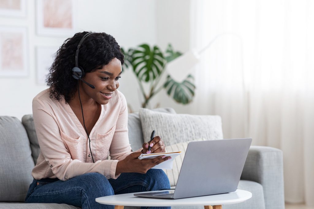 Online Education. Young Black Female Studying Online From Home With Laptop Computer And Headset And Taking Notes To Notepad, Enjoying Distance Learning, Watching Webinar In Internet, Copy Space