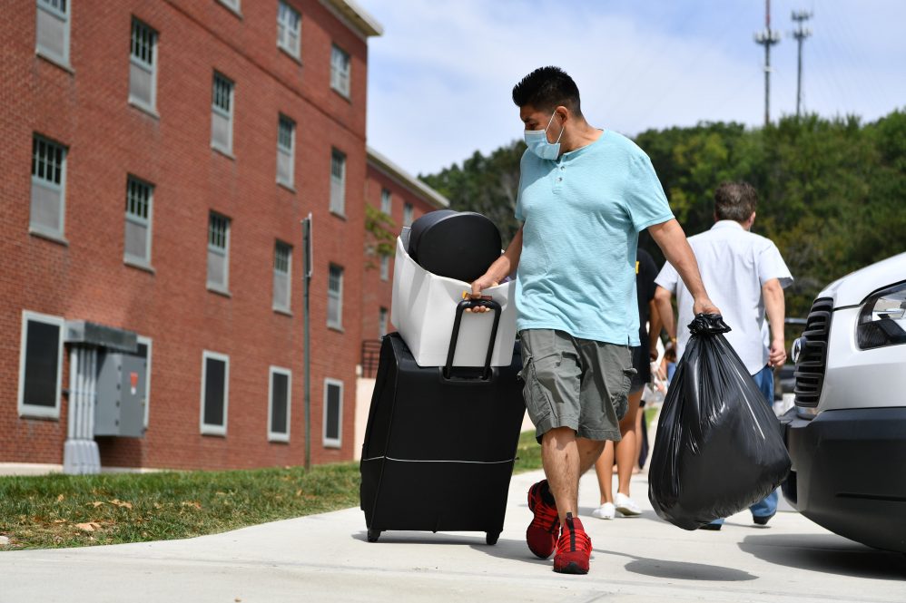 A student with a facemask carrying a suitcase behind him outside a residence hall, preparing to move in.