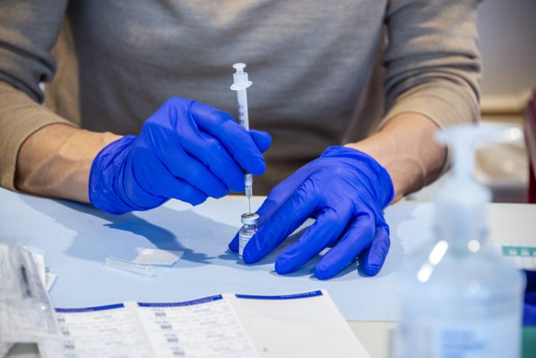A person in blue medical gloves using a syringe to draw COVID-19 vaccine from a vial.
