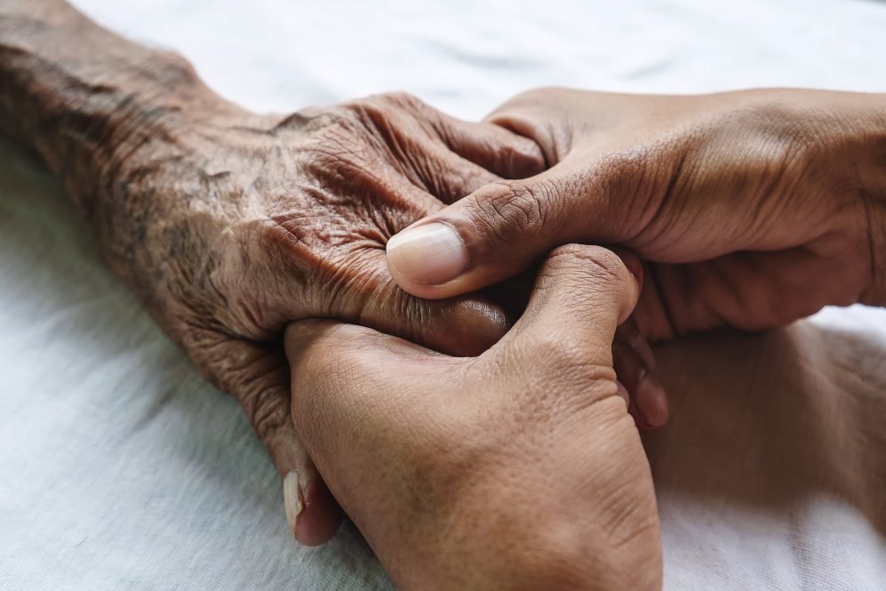A younger man holds an older man's hands on a bed. UConn Health researchers have received a grant to investigate new methods of addressing health complications that come with aging.