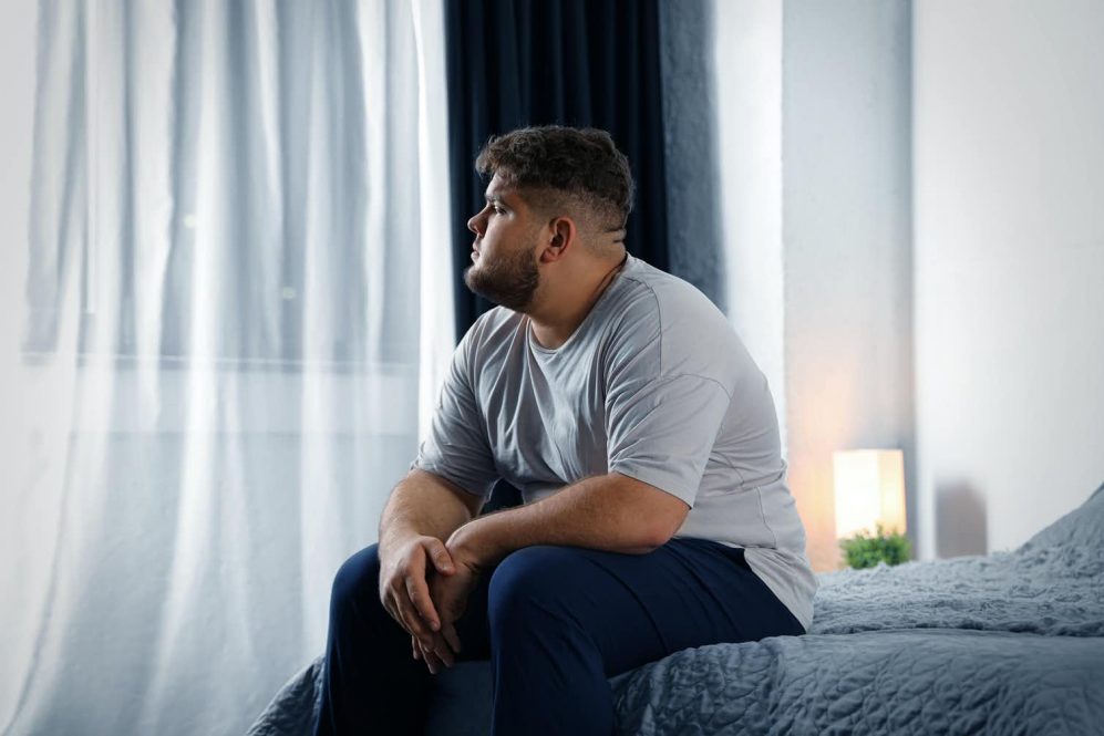 An overweight young man sits on a bed, gazing out the window with an air of melancholy. People with type 2 diabetes face stigma that can adversely affect their health.