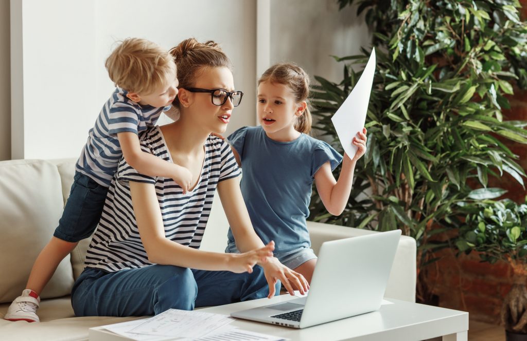 Tired young mother sitting on sofa and working with laptop and documents while little kids having fun and making noise.