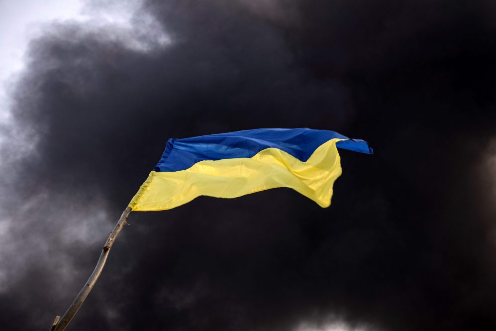 A Ukrainian national flag waves while smoke rises after Russian attacks hit a fuel storage facility in the city of Kalynivka, on March 25, 2022.