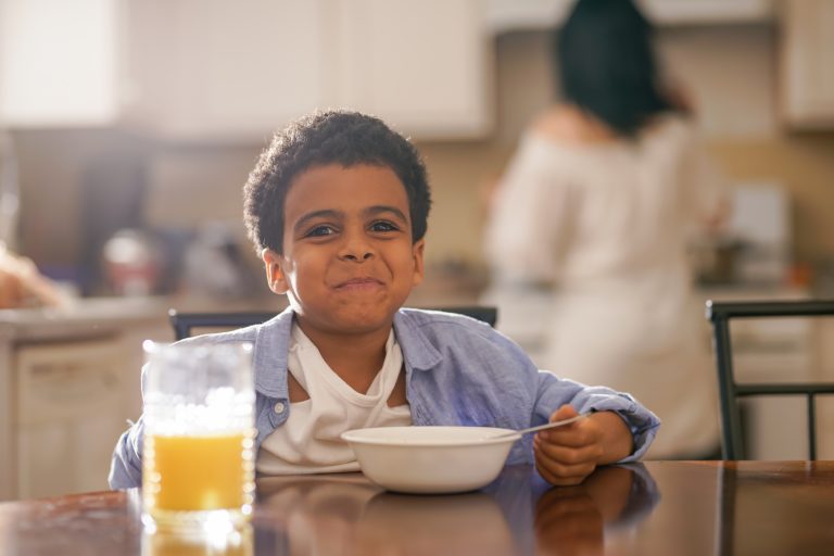 A young child eats cereal at the breakfast table in a kitchen with morning light streaming in behind him.