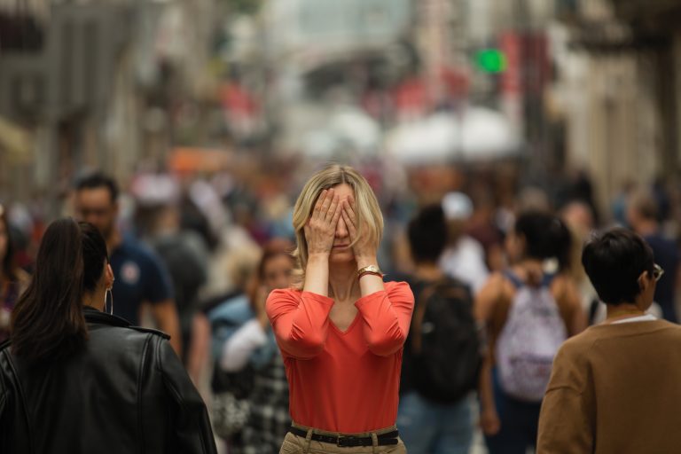 Sad depressed woman covers her eyes with her hands surrounded by people walking in crowded street. Panic attack in public place.