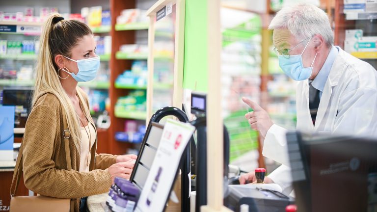Senior pharmacist dealing with a customer, both of them wearing masks due to coronavirus.