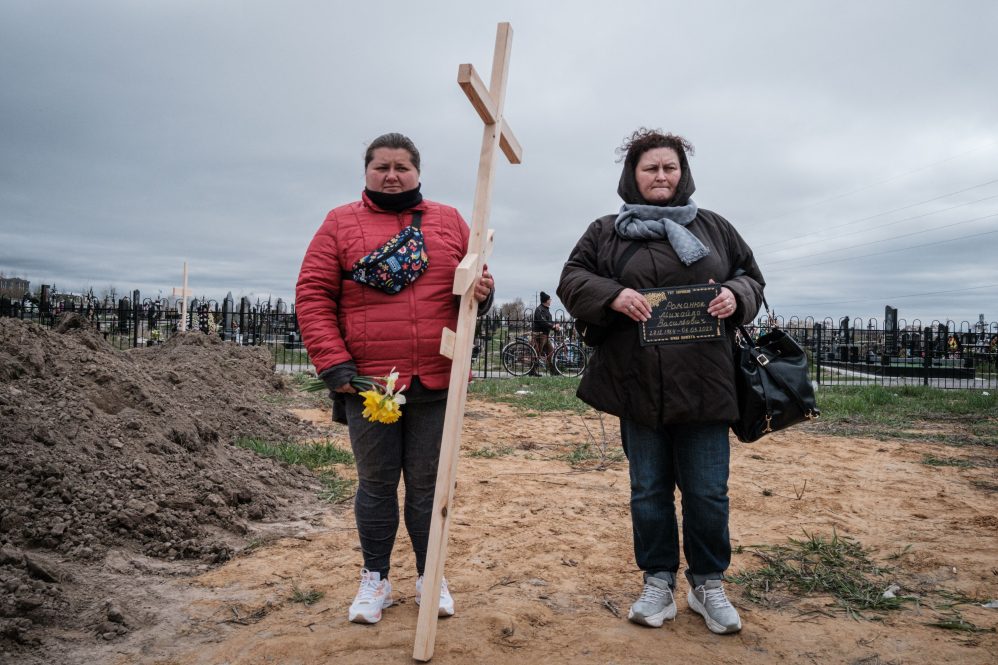 Two women stand on a dirt road, one carrying a wooden cross. They are waiting to bury a relative killed in the Russian invasion of Ukraine.