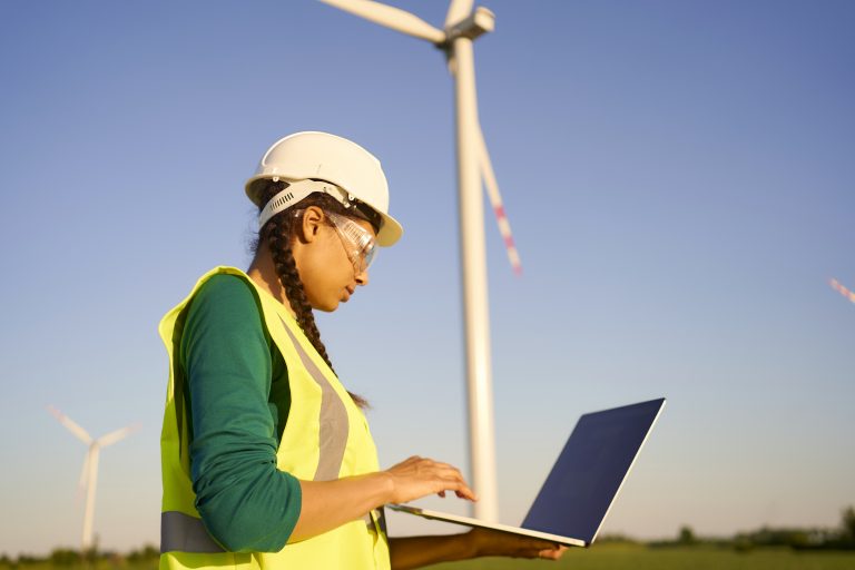 Female engineer wearing hard hat and reflective green jacket standing with laptop against wind turbine and setting it up.