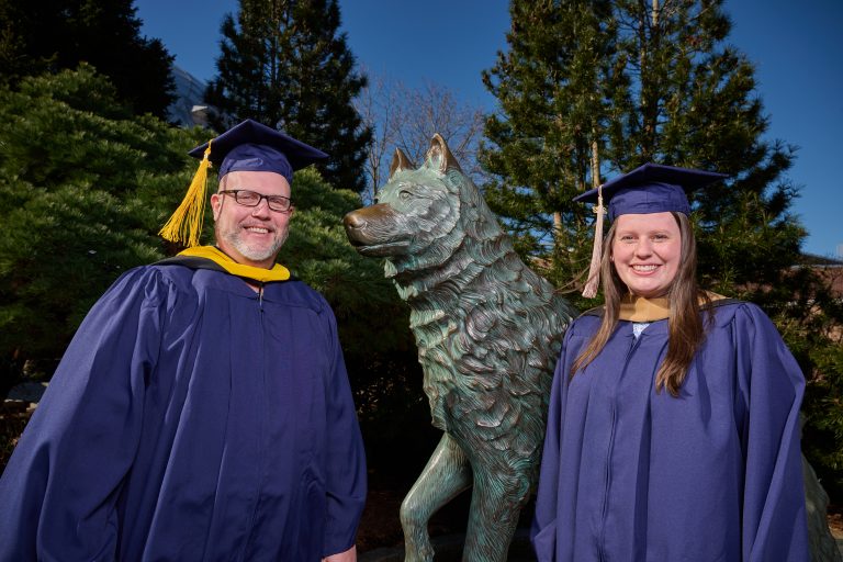 Kyle Muncy '92 (CLAS) '22 MS, director of brand partnerships and trademark management, left, and his daughter Taylor Muncy '19 (CLAS) '22 MBA, wear their caps and gowns while standing next to the Jonathan the Husky statue on April 11, 2022.