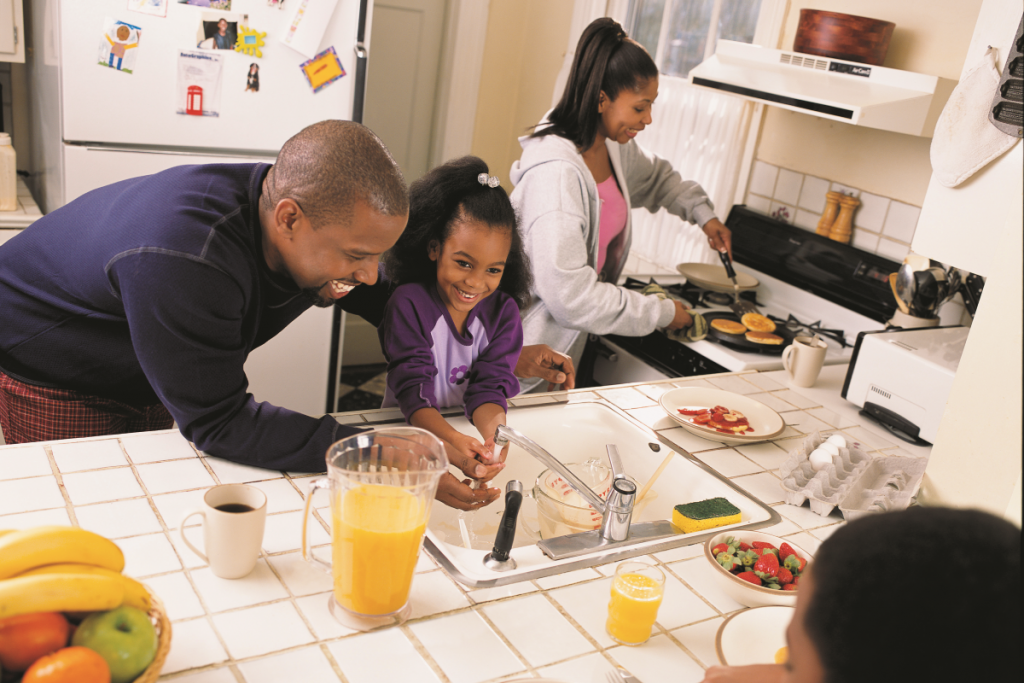 A man and his daughter wash their hands while a woman prepares breakfast in the background
