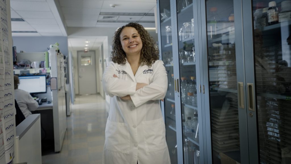 Jenna Bartley, a UConn researcher on aging and health, stands in a hallway at UConn Health, wearing a white lab coat.