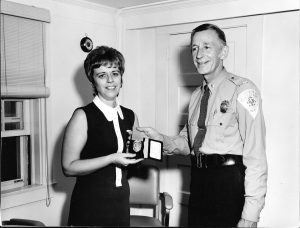 A black and white photo of a woman in civilian clothes and a man in a police uniform. She is holding a wallet with a badge.