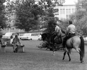 A black and white photo that shows two UConn police officers on horseback, chatting with two students sitting on a bench.