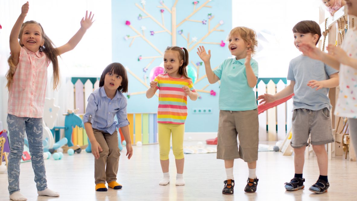 Cheerful kids stand semicircle on floor in kindergarten or daycare centre. Preschoolers have fun indoors, playing games.