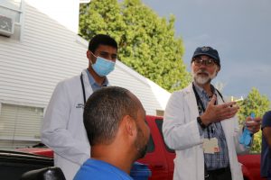 A UConn Health doctor, in beard and white coat, speaks with a man in a Hartford neighborhood while a masked medical student stands next to him.