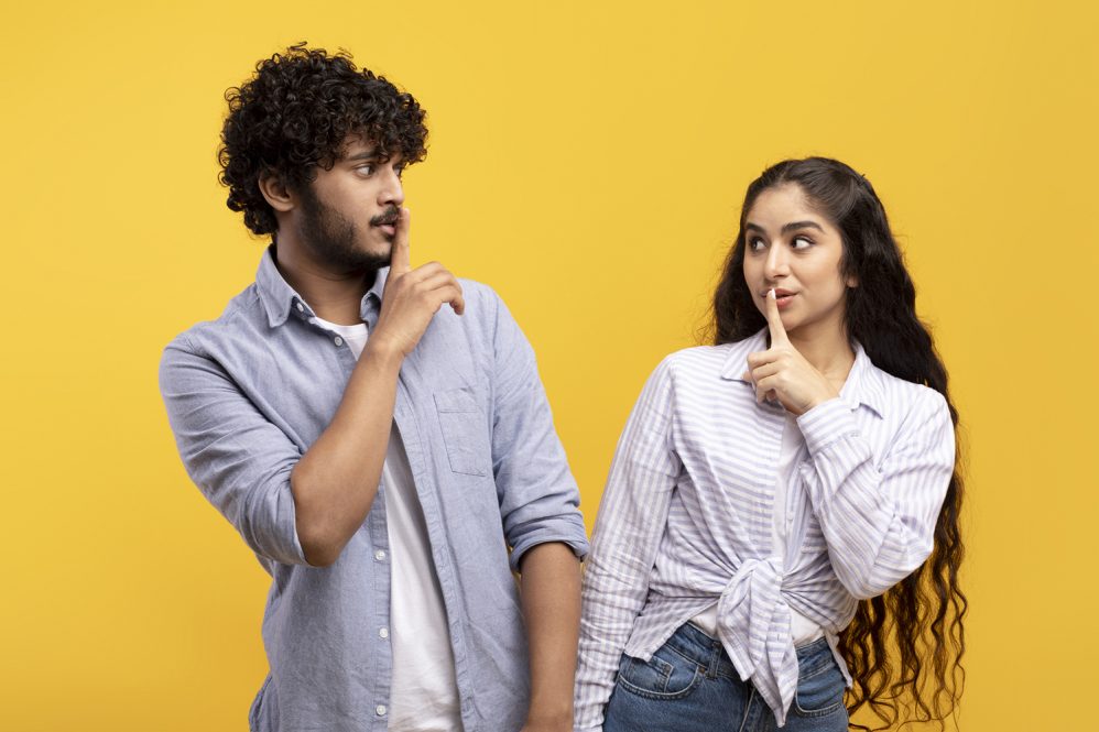 A young couple standing against a yellow background make the "shush" finger gesture at one another.