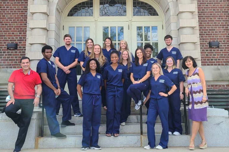 A group of students in scrubs stands on the steps of a campus building.