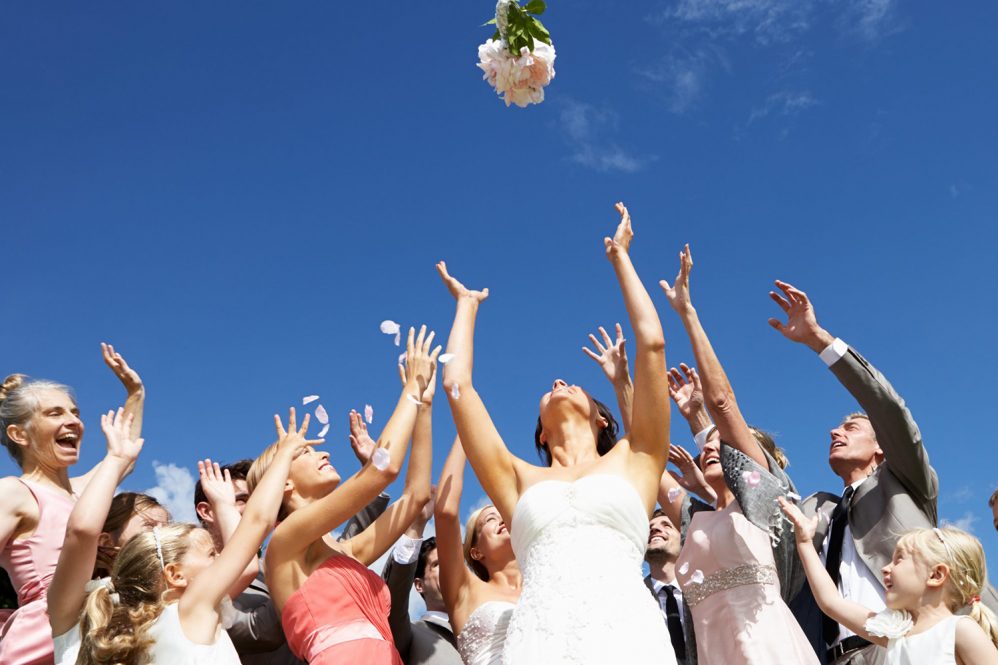 A bride throws a bouquet of flowers over her shoulder as guests stretch out their arms to catch it.