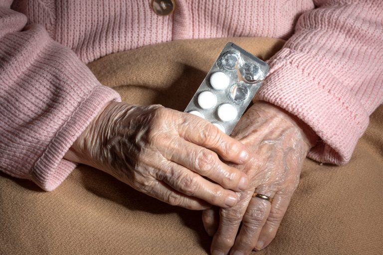 The hands of an elderly woman clutch a blister pack of medication, with three tablets missing.