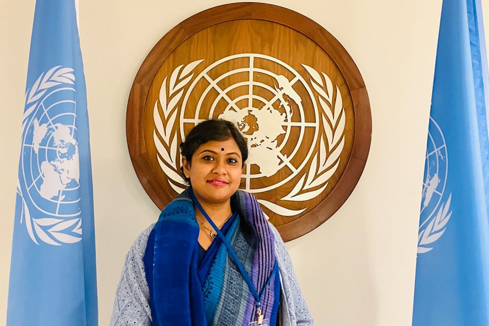 Photo of Rianka Roy standing in front of the UN logo