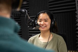 Andrea Li wears headphones while sitting in front of a microphone in a recording studio.