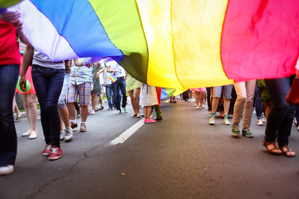 A Pride flag being carried in a parade by people of different races and ethnic backgrounds.