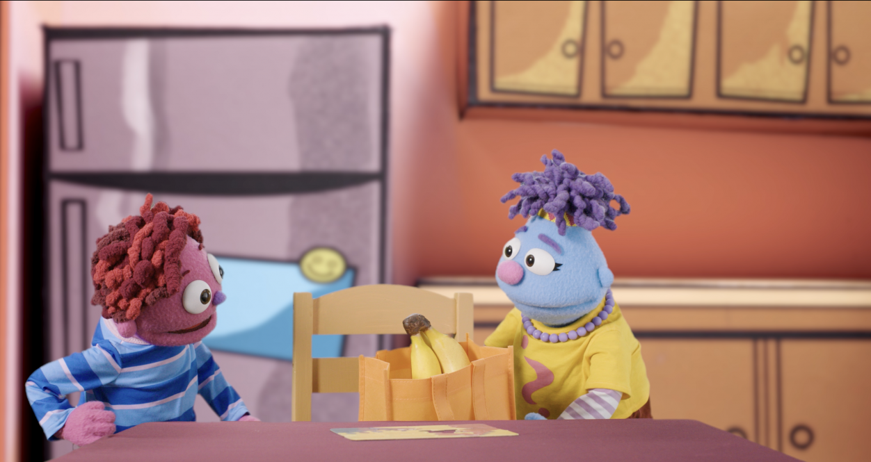 Two puppet characters discuss a paper sack filled with bananas in front of a colorful painted kitchen background in this video still from Feel Your Best Self's "Be a Kind Helper."