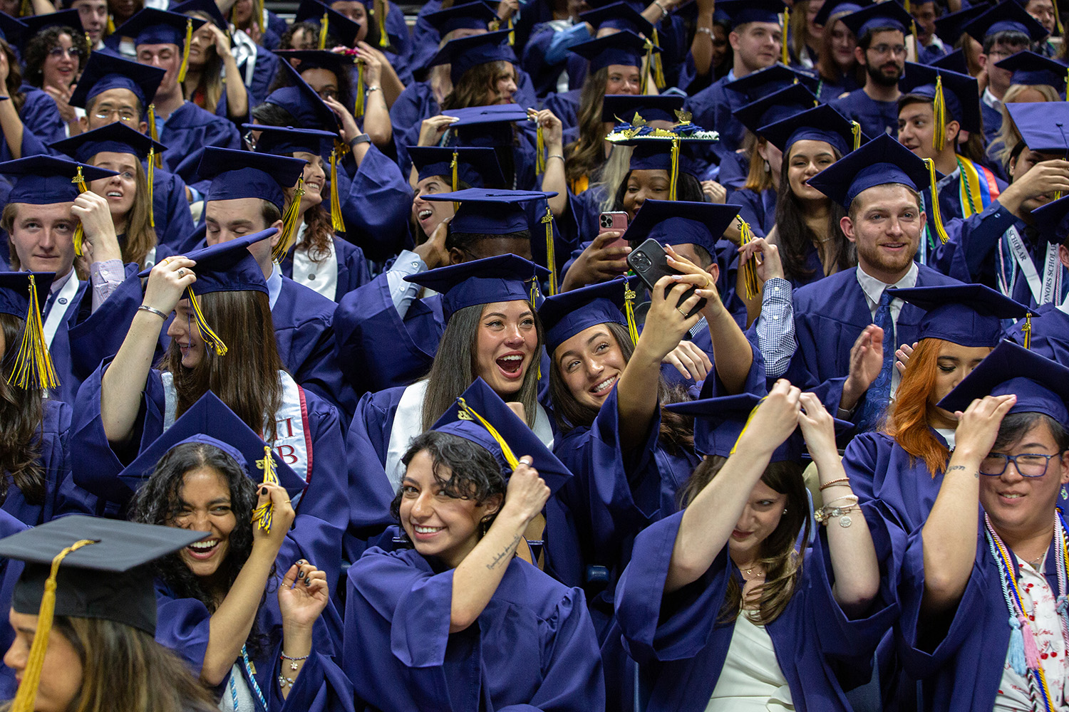 Students in full regalia move their tassels and pose for selfies during commencement.
