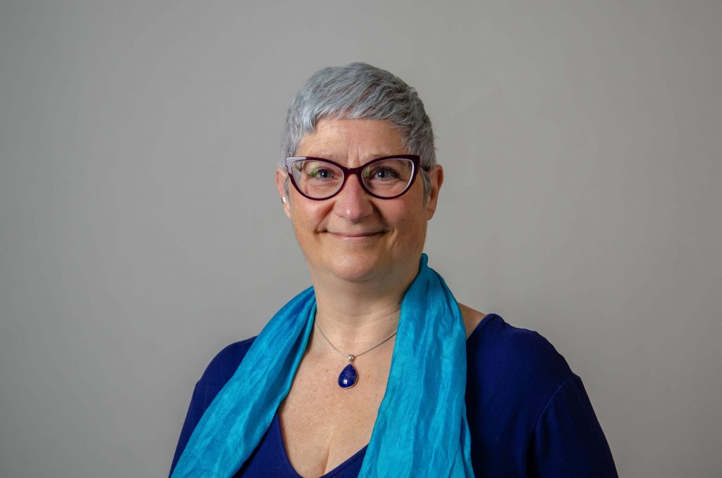Marie Coppola, wearing a blue scarf, necklace, and purple glasses, smiles at the camera.