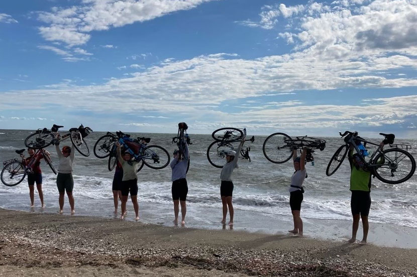 Eight cyclists hoisting their bikes with their feet in the water on the ocean shore