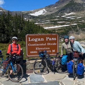 Three cyclists in front of Logan Pass sign, mountains in background