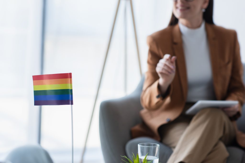 A Pride flag in the foreground, with a mental health professional seated in the distance.