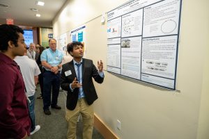 A young researcher wearing khakis, a blazer, and necktie presents a poster to audience members.