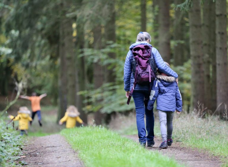 In the foreground, an older woman puts her arm around a young child as they walk together down a woodland trail. More children can be seen running and playing up ahead on the trail.