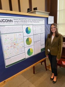 Lucie Lopez stands to the right of a research poster, whose title is "Grocery Shopping Near and Far: How Being Food Insecure and Living in a Food Desert Can Impact Shopping Habits."