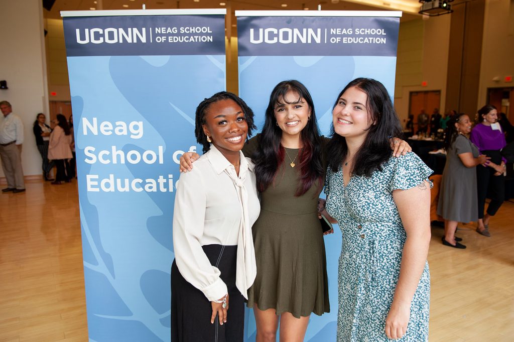 Three smiling females stand in front of blue banner with letters "Neag School of Education."