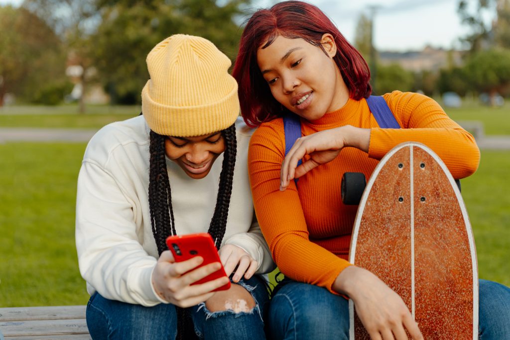 Two young women of color look at a smart phone while sitting together in a park.