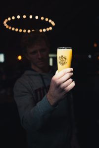 A student in shadow holds a glass of beer in the light.