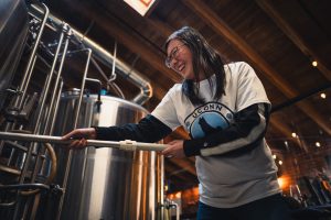 A student with a UConn shirt stirs a combination of milled grains and warm water in a large brewery vat.