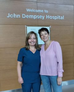Two women standing in front of "Welcome to John Dempsey Hospital" sign