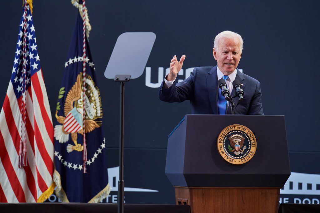 US President Joe Biden stands behind a podium delivering a speech. The UConn and Dodd Center logos are visible on the wall behind him, and an American flag and flag with the presidential seal are visible to his right.