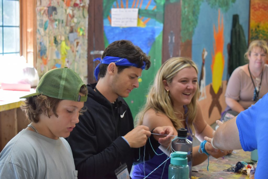Three people sit in a room with a colorful mural of a campfire in the background, laughing and tying friendship bracelets.