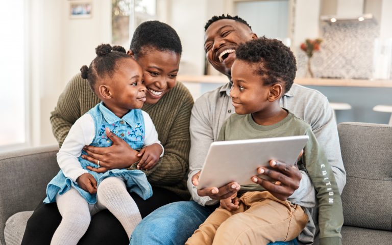 Two parents and two young children sit together on a sofa, laughing about what they are seeing on a digital tablet.