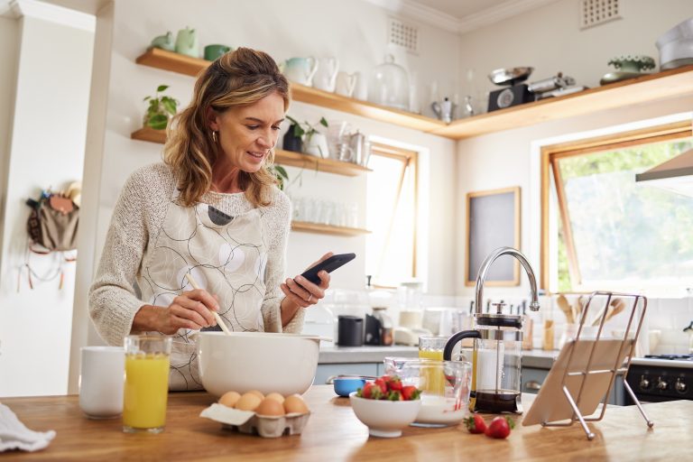 A woman cooks in her kitchen while consulting her phone for information on healthy choices.