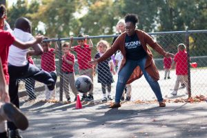 A female college student plays soccer with elementary school students outside on a blacktop.