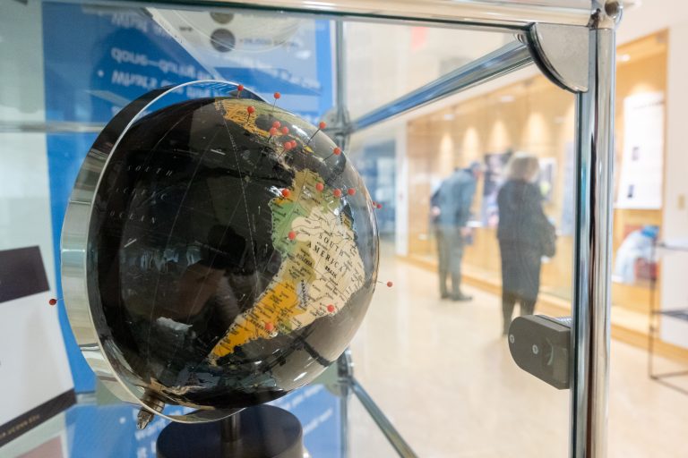 A globe in a museum display case, with red pins marking locations.