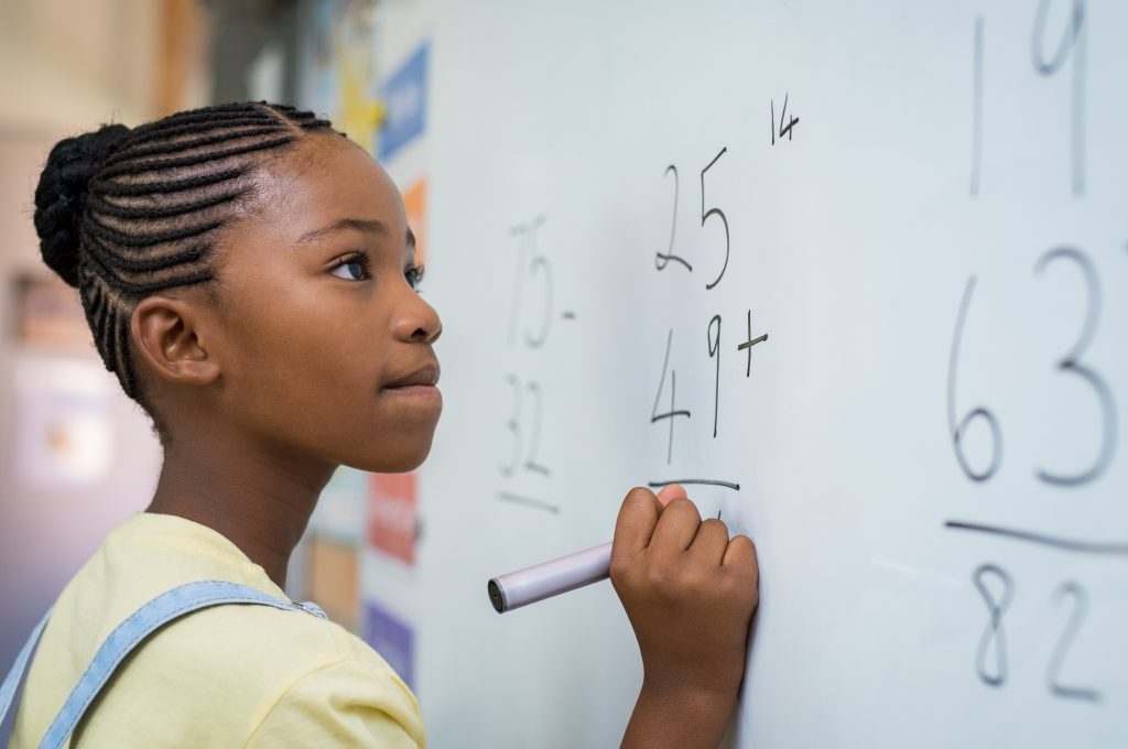 Black schoolgirl solving addition sum on white board with marker pen.