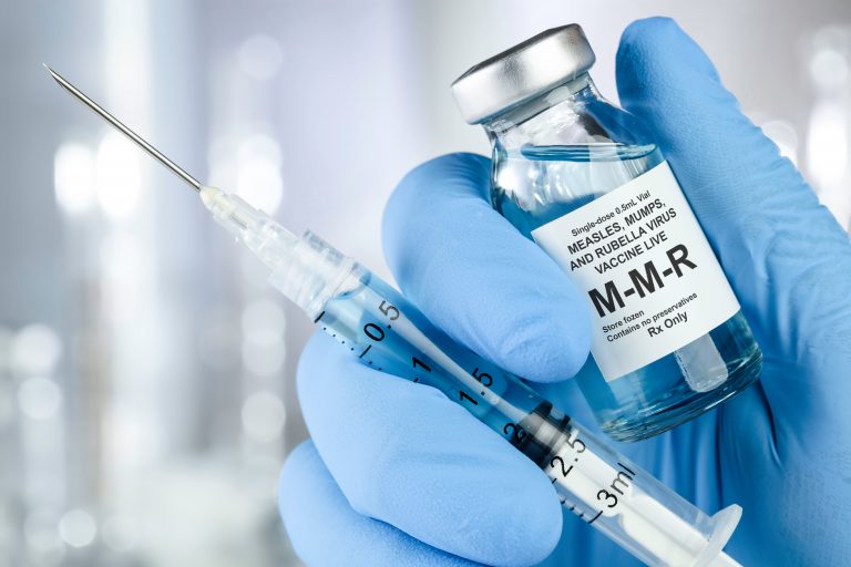 A hand clad in blue surgical glove holds a syringe and vial of MMR vaccine.