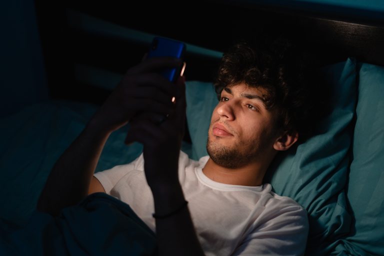 A young man in bed at night, his face illuminated by the glow of the cell phone he holds in his hands.