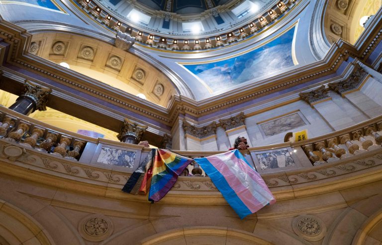 A Pride flag and a trans flag hang side by side over a railing inside a state capitol building.